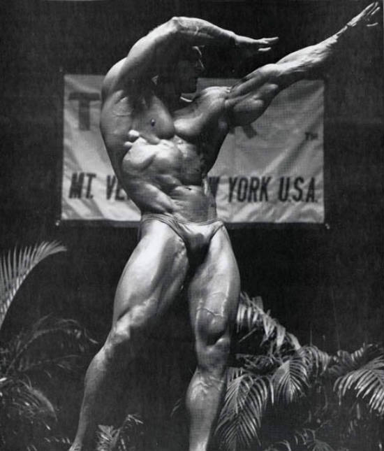 Frank Richards posing in a archers position, showing his big triceps, abs and large quads 