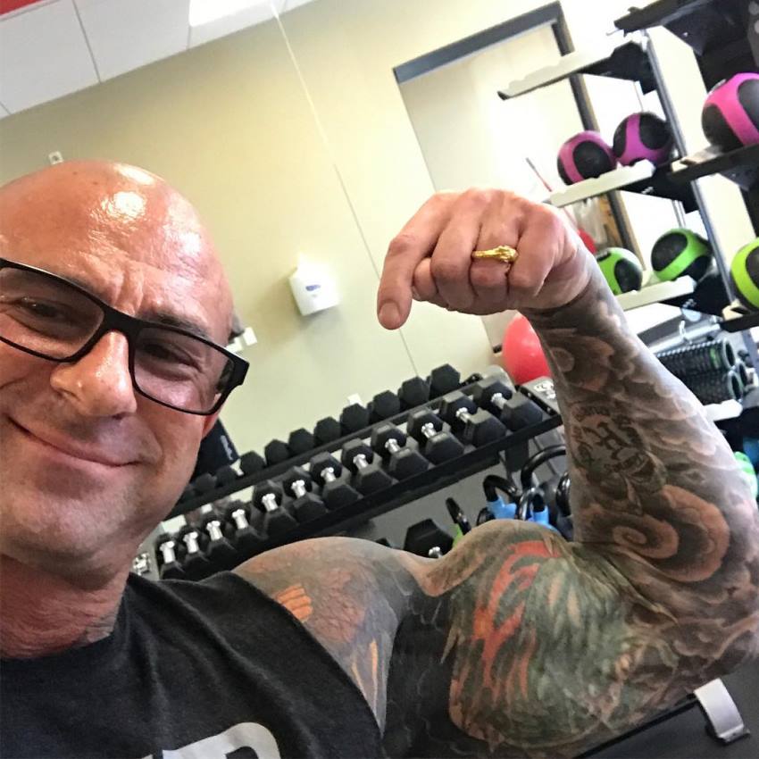 Jim Stoppani wearing glasses, and doing a biceps-flex selfie in a gym, smiling at the camera