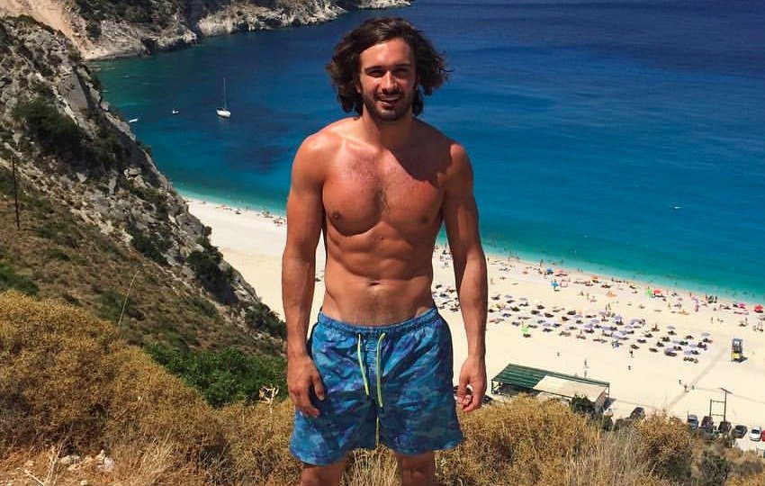 Joe Wicks on a hill which overlooks a beach, flexing his lean abs and chest