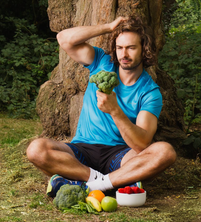 Joe Wicks sitting besides a tree with broccoli in his hand, and other vegetables and fruits on the ground, having one of his hands in the air, and thinking about something