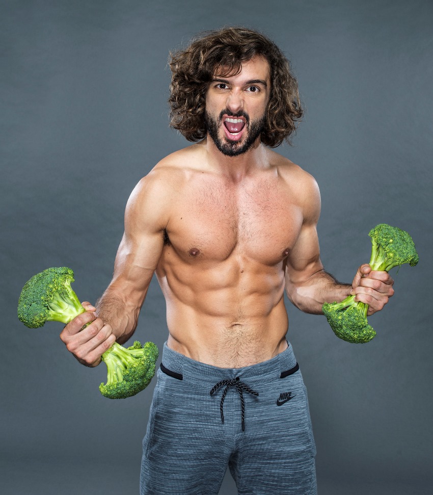 Shirtless Joe Wicks yelling at the camera with a grimace on his face, while holding broccoli in each of his hands