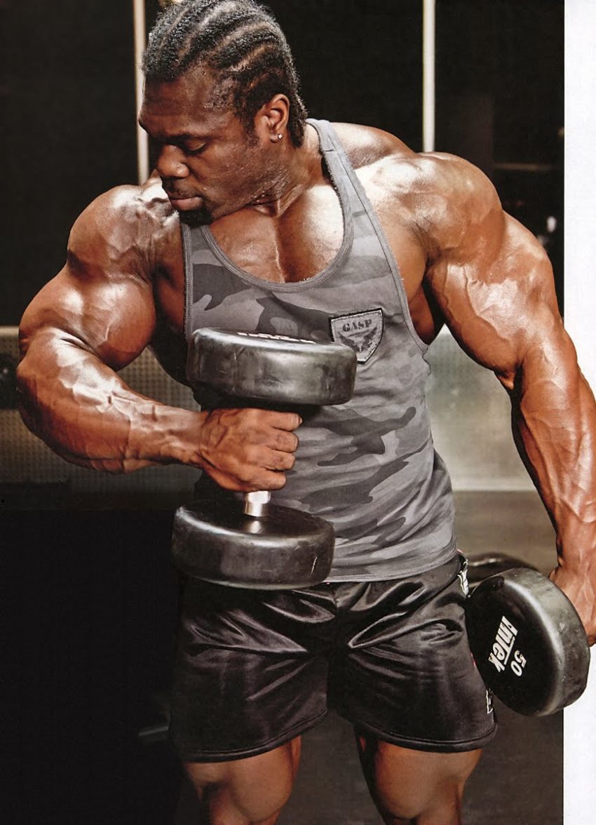 Lionel Beyeke in a grey camouflaged tank top, doing hammer curls with his big muscular arms