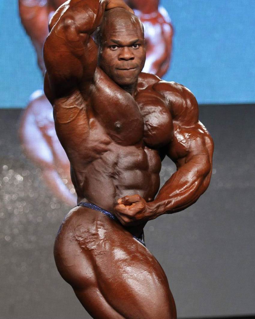 Lionel Beyeke in a side most muscular pose, flexing his ripped abs, glutes, legs. chest, and arms