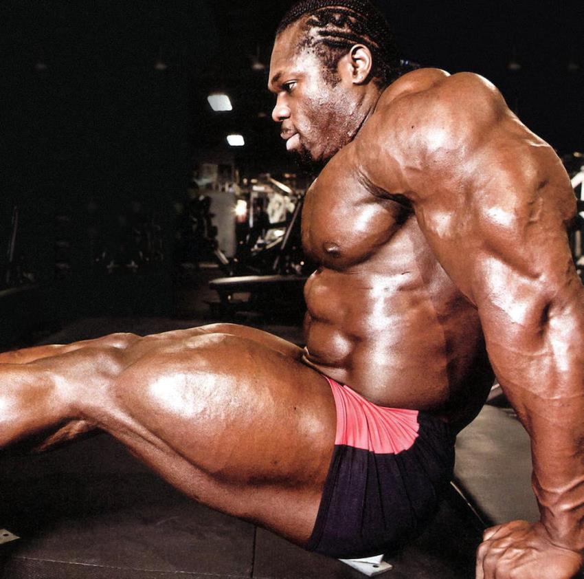 Lionel Beyeke doing triceps dips, while also flexing his entire arm muscles, as well as legs and abs