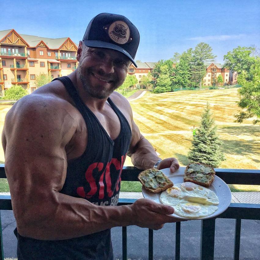 Marc Lobliner on balcony which looks over a beautiful nature scenery, holding a plate with food in his hand, smiling at the camera, and showing her big muscular arm