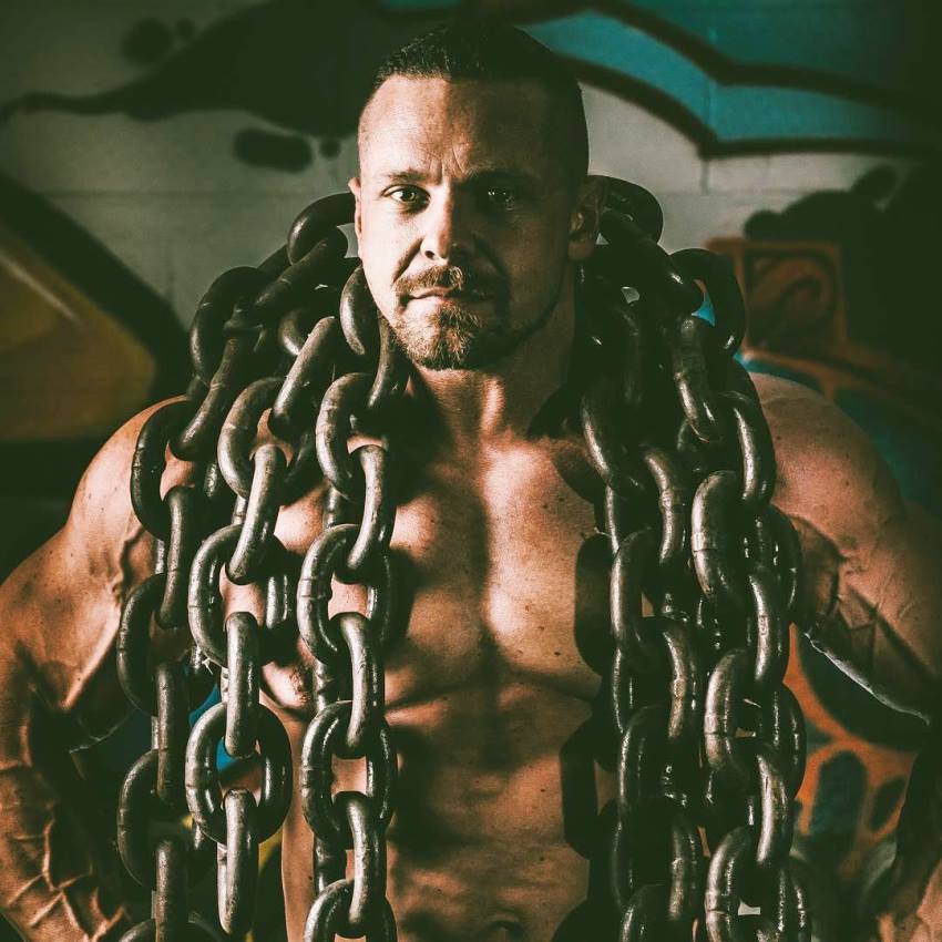 Shirtless Marc Lobliner with many heavy chains around his neck, looking straight into the camera