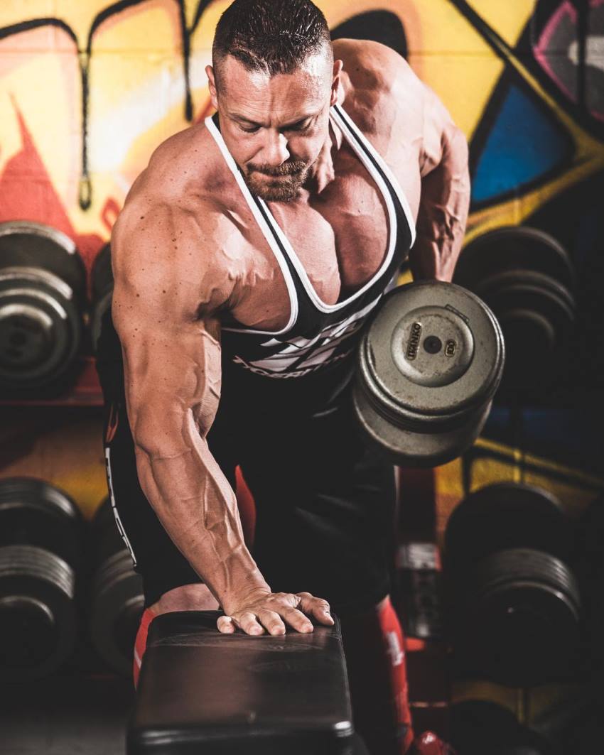 Marc Lobliner in a tank top, doing single-hand dumbbell-bent over rows on a flat bench