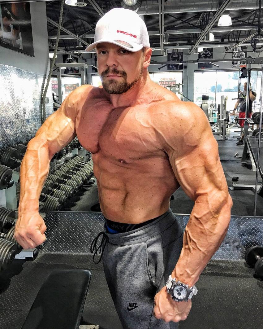 Marc Lobliner doing a shirtless side pose in a gym, showing his vascular and ripped arms, shoulders, pecs, and abs