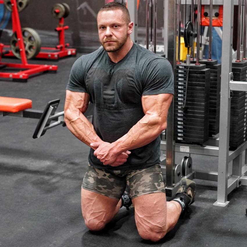 Marc Lobliner kneeling on a gym floor in front of a cable machine, as he looks into the camera, displaying his vascular legs and arms