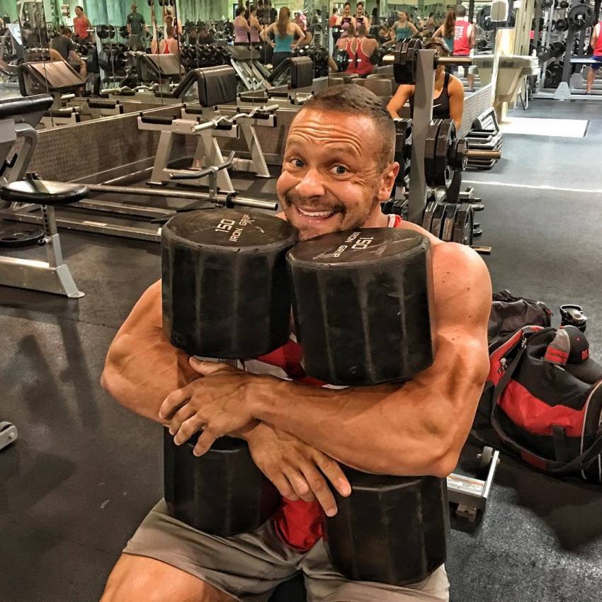 Marc Lobliner sitting on a flat bench, holding two extremely heavy dumbbells, as he hugs them, and smiles excitedly at the camera