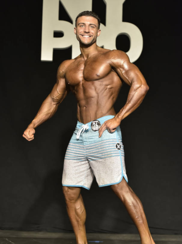 Matt Acton showing his full body at a competition, tensing his vascular arms and chest