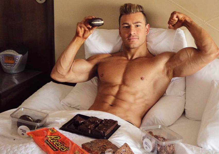 Maxime Parisi eating cheat food and showing his ripped abs and large bicep in bed 