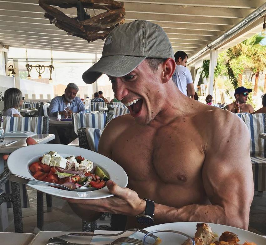 Maxx Chewning sitting shirtless in an outdoor restaurant, looking at a plate full of food with an excited expression on his face