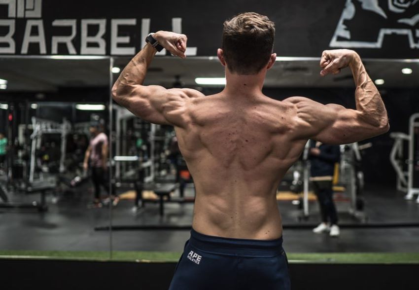 Maxx Chewning in a shirtless back double biceps pose in a gym, showing his shredded back, arms, and shoulders