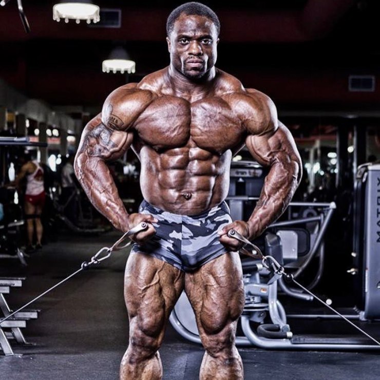 Michael Lockett holding two cables in his arms in the gym, being shirtless, showing his massive and shredded physique to the camera