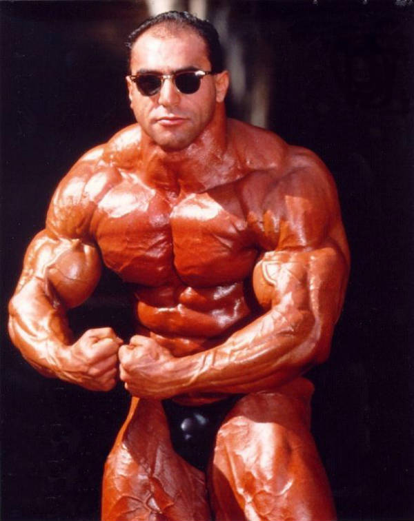 Nasser El Sonbaty tensing his arms whiel wearing sunglasses, showing off his massive delts, traps and biceps