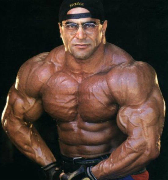 Nasser El Sonbaty tensing his biveps while wearing a baseball cap and weight lifting gloves