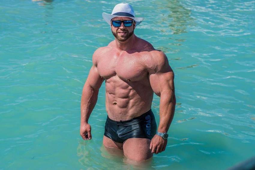 Petar Klancir standing in a pool with a white hat and blue shades on, showing off his massive and lean body