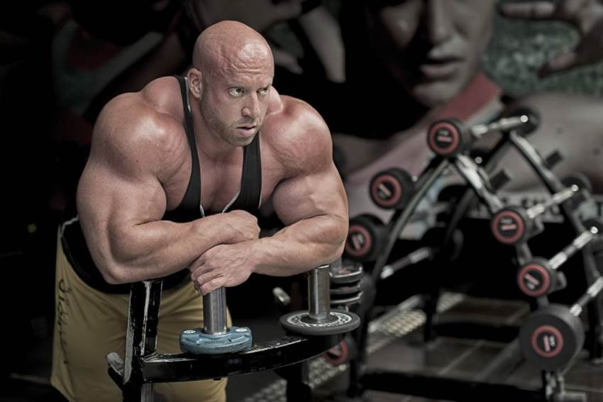 Petar Klancir leaning against a gym machine, looking at the distance, with his arms and shoulders looking massive and round