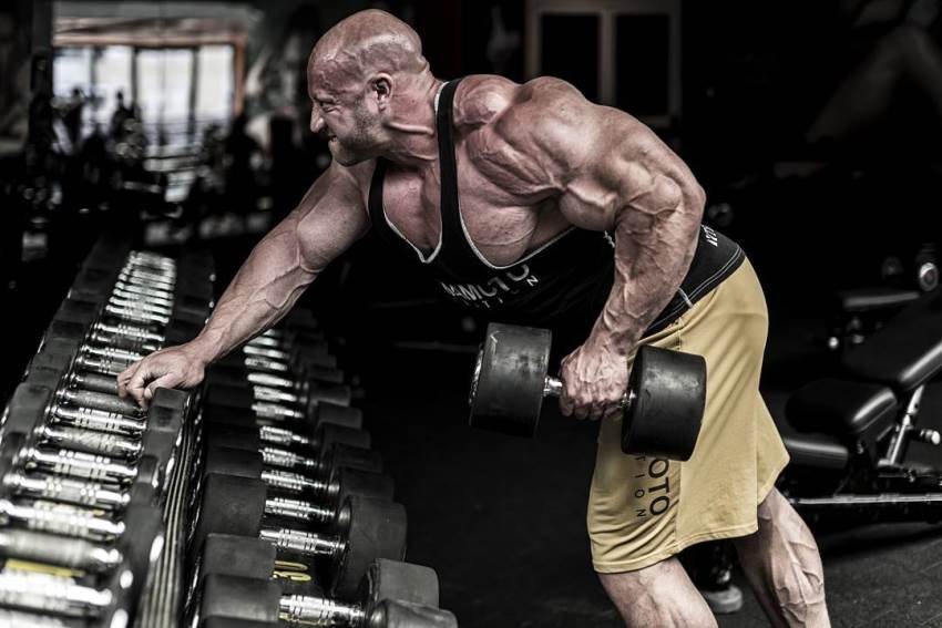 Petar Klancir leaning against dumbbell rack, doing single-arm bent over rows