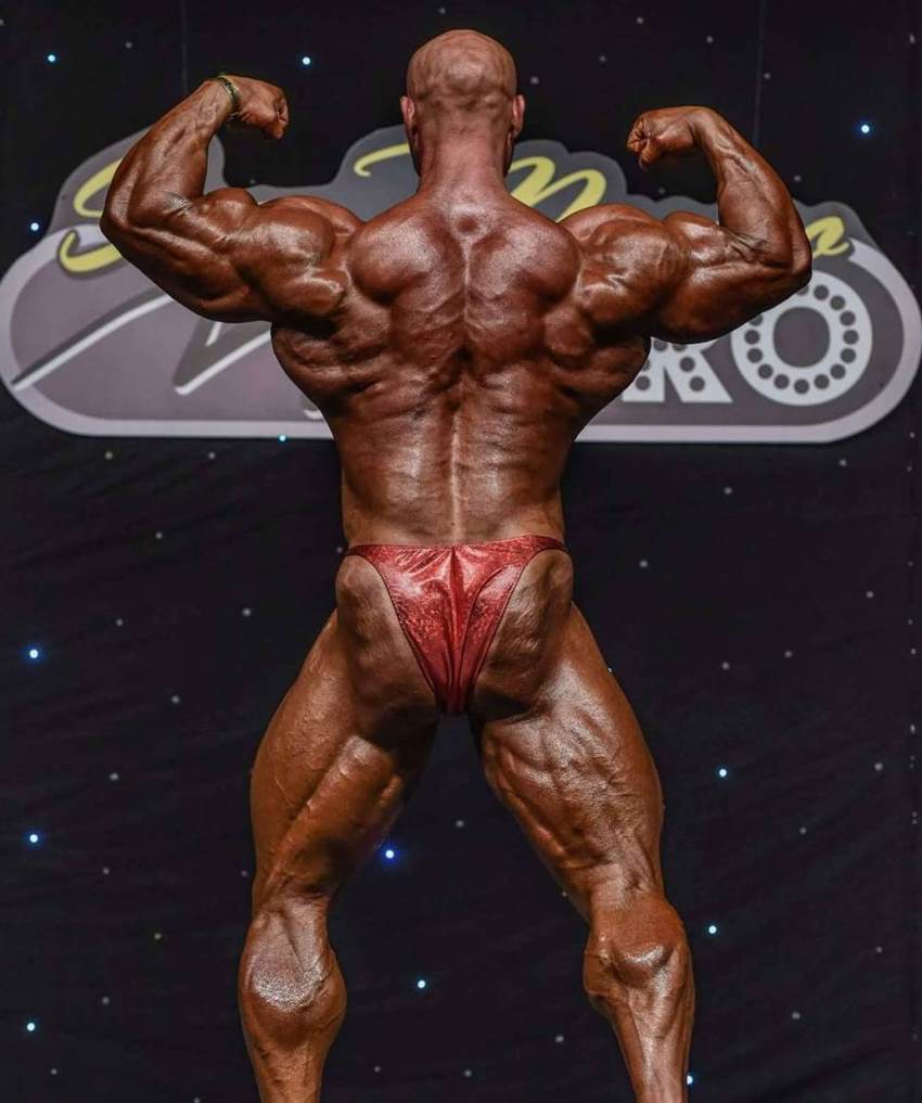 Petar Klancir on a bodybuilding stage in red trunks, holding an impressive back double biceps pose