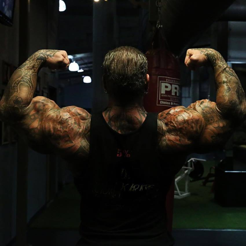 Rich Piana in a back double biceps pose in a black tank top, flexing in the dark, showing his massive tattooed arms and shoulders