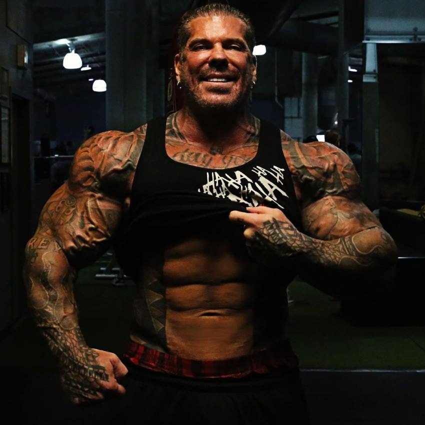 Rich Piana smiling as he raises his tank top, showing his impressive ripped abs, all while being massive and muscular