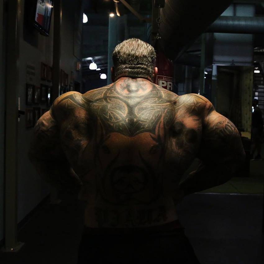 Rich Piana shirtless in the dark, showing his massive tattooed back in a rear lat spread pose