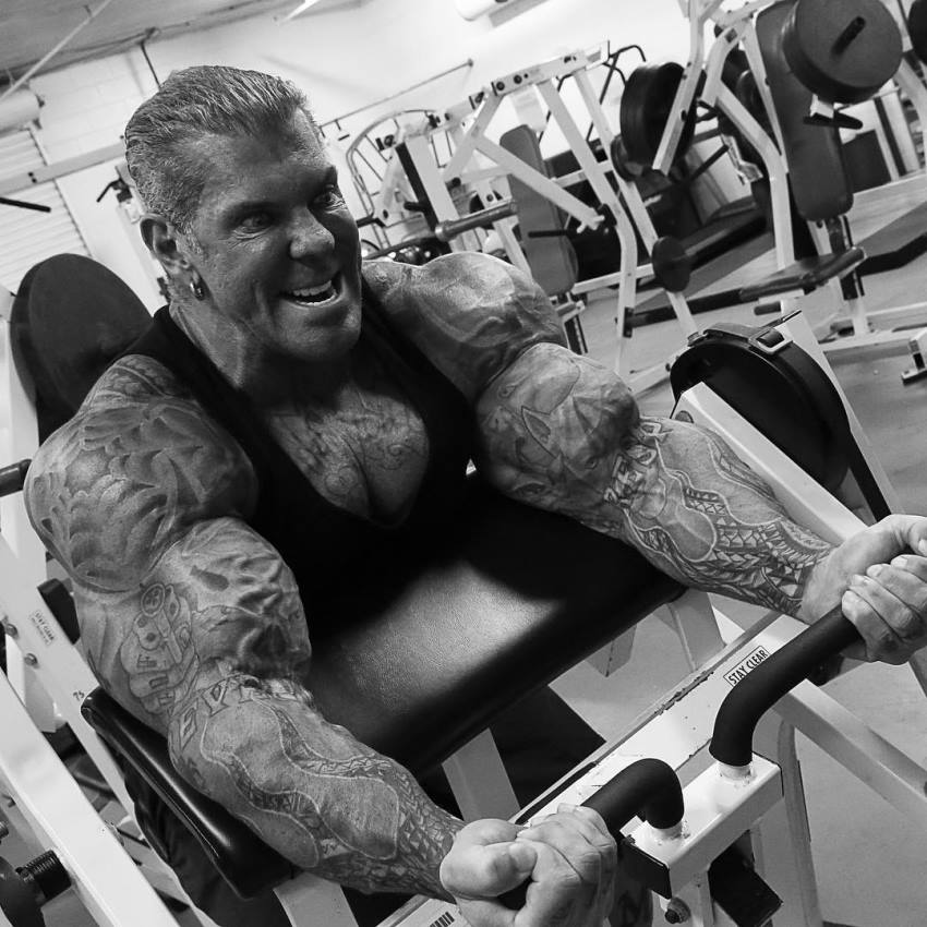 Rich Piana doing biceps curls on a machine in a gym, with a pained grimace, showing his incredible arms