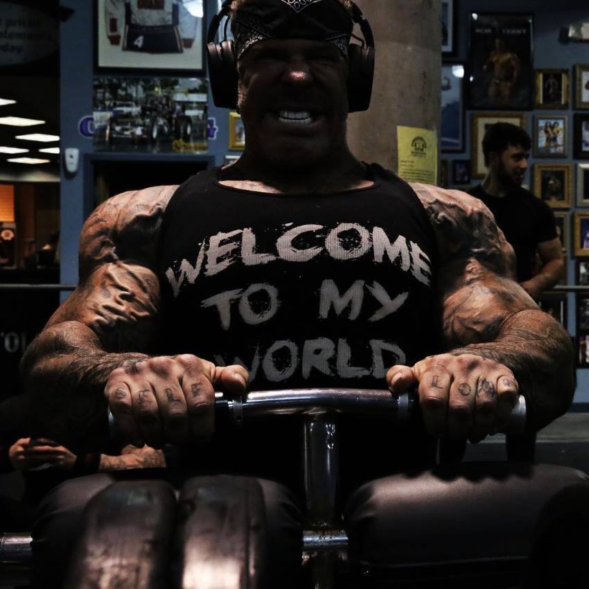 Rich Piana doing seated calf raises in a black tank top and headphones over his head, having a pained face expression as he does the exercise