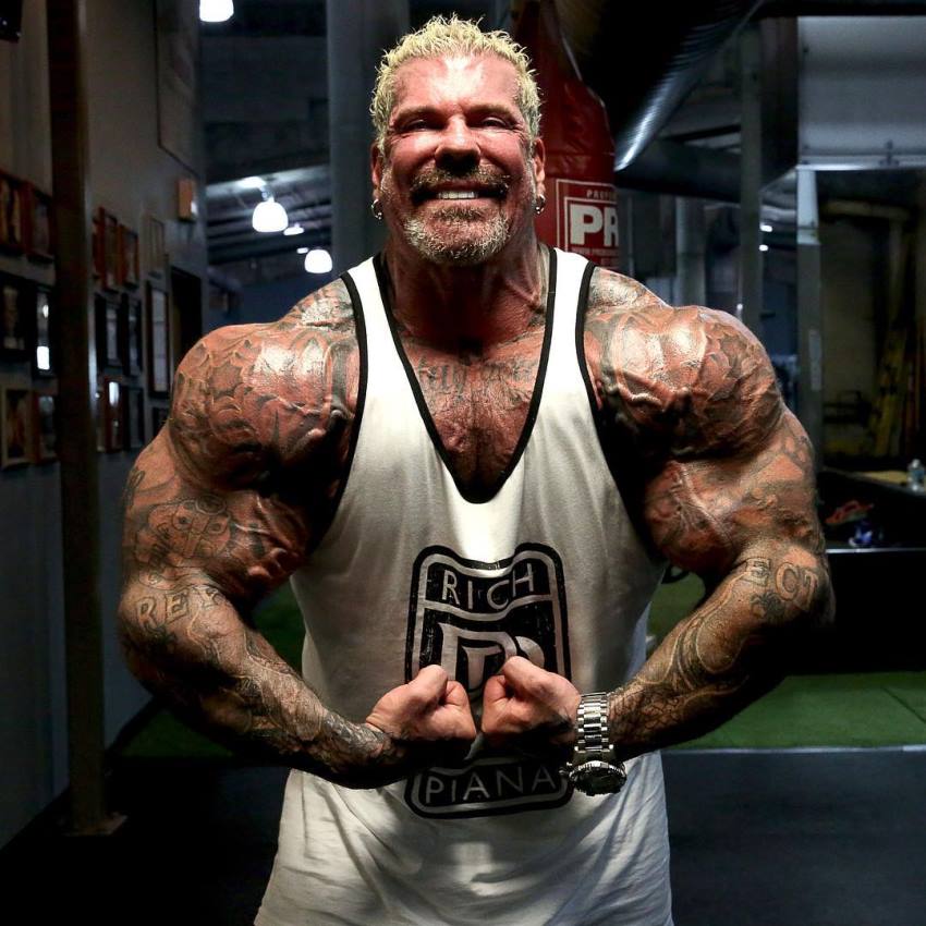Rich Piana in a white tank top with black outlines, flexing his arms for the camera, as he stands under the perfect downlighting which accentuates his physique