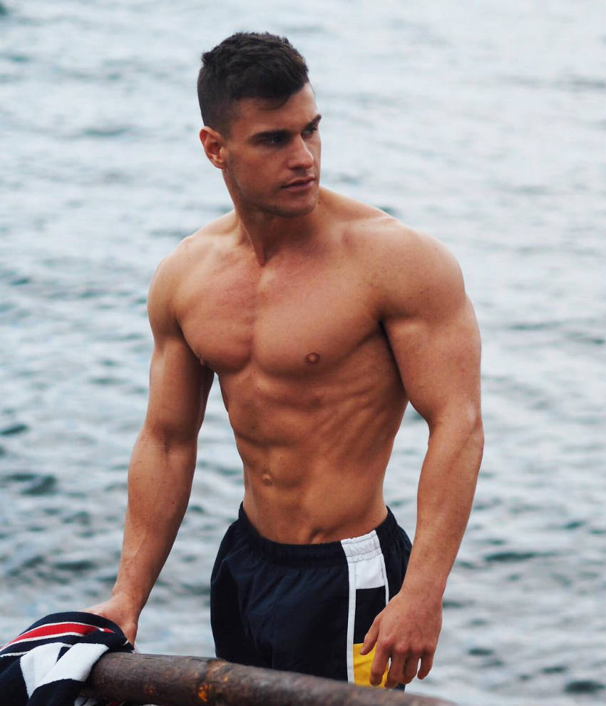 Rob Lipsett standing next to the ocean, showing his ripped abs, obliques and large delts