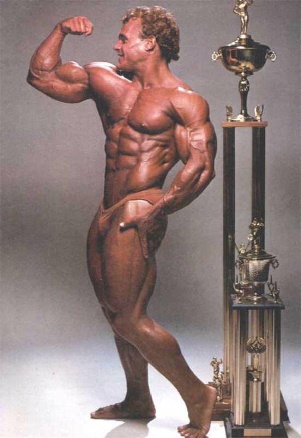 Tim Belknap standing next to a competitions trophy, tensing his bicep and showing his muscular body 