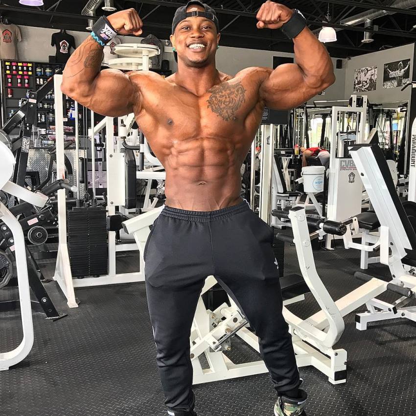 Brandon Hendrickson doing a shirtless front double biceps pose in the gym, looking muscular and lean