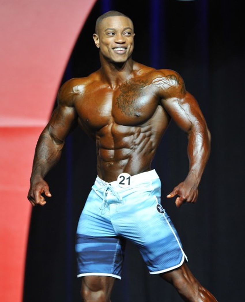 Brandon Hendrickson confidently posing on the Men's Physique Olympia stage, having a big smile on his face, as she shows his awesome and conditioned physique to the audience