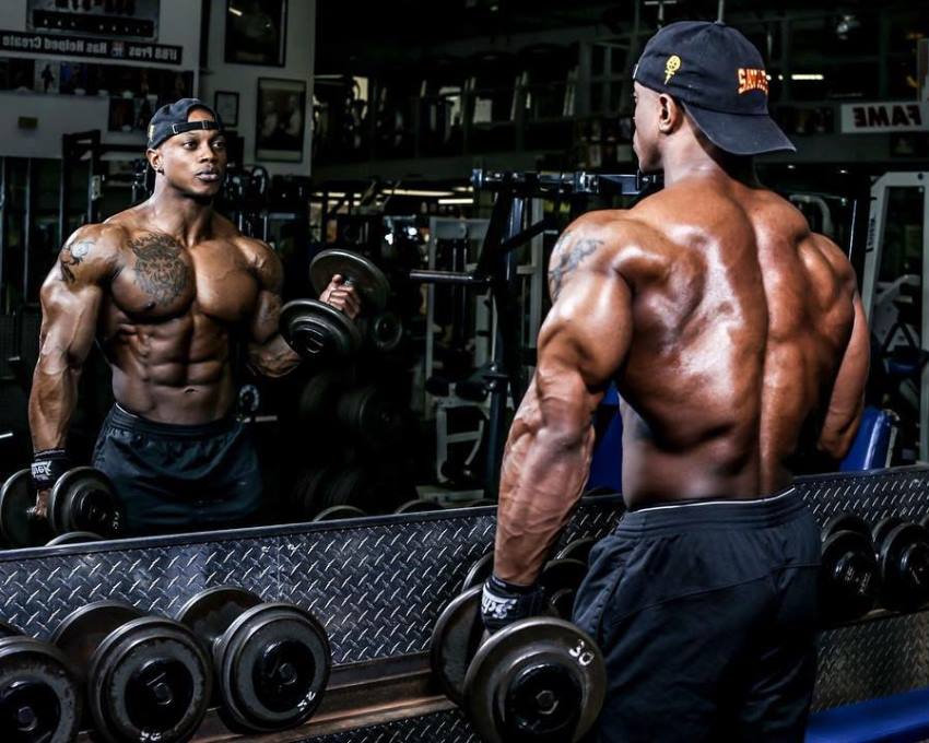 Brandon Hendrickson looking at himself in the gym mirror while doing biceps curls, looking ripped and muscular