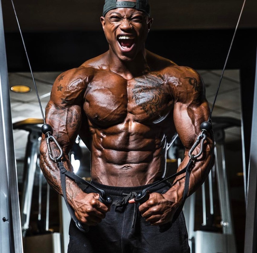 Shirtless Brandon Hendrickson doing cable crossovers in the gym, showing his ripped body, having pained expression on his face