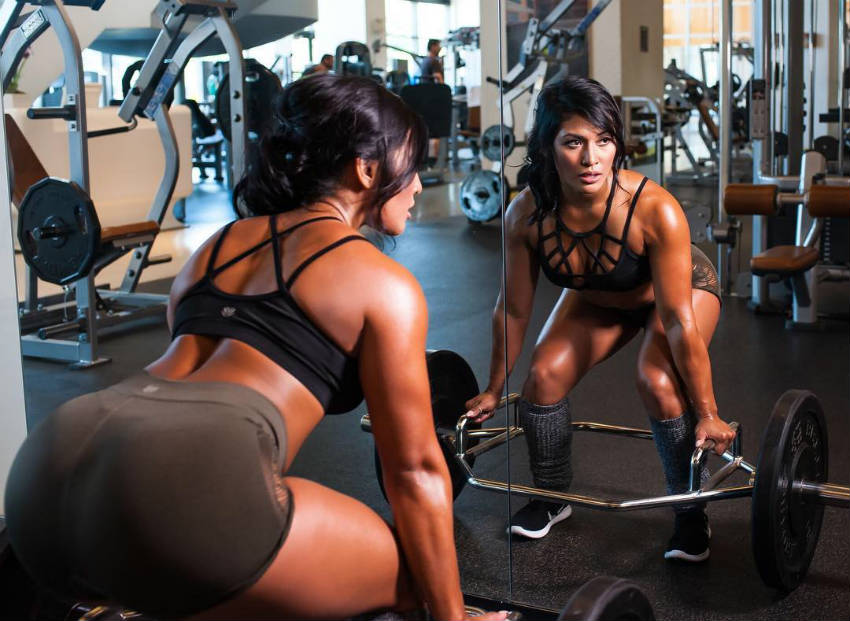 Genevieve Ava completng a deadlift, looking into the mirror and showing of her glutes and abs