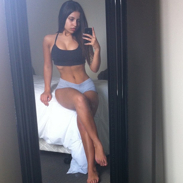 Gina Savage sitting on a bed and taking a selfie of her lean legs, abs, and arms