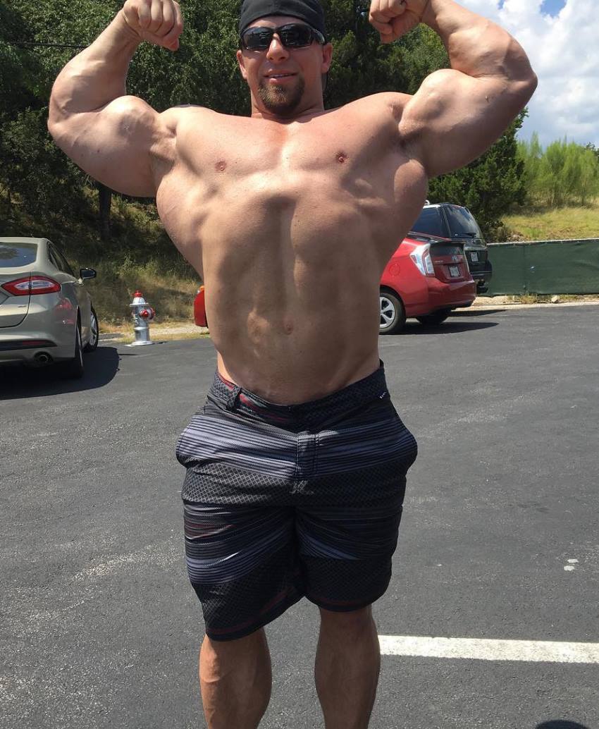 John Jewett in a shirtless front double biceps pose in an outdoor parking lot, showing his lean and big physique