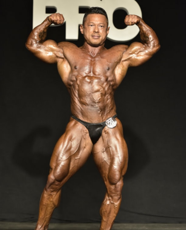 Leonardo Pacheco performing a front double biceps pose on the stage, looking muscular and ripped