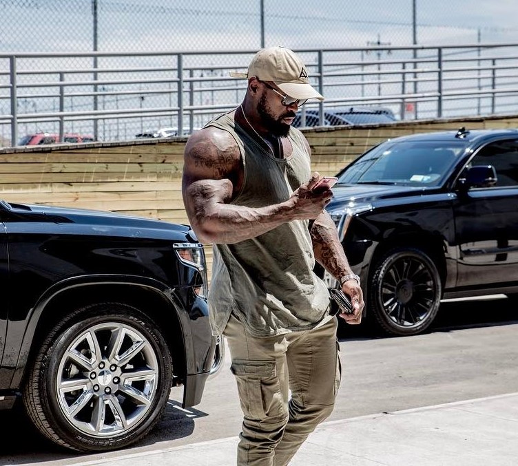 Mike Rashid outside in green tanktop, looking at his mobile phone as he's walking out of his car, his arms looking muscular and ripped
