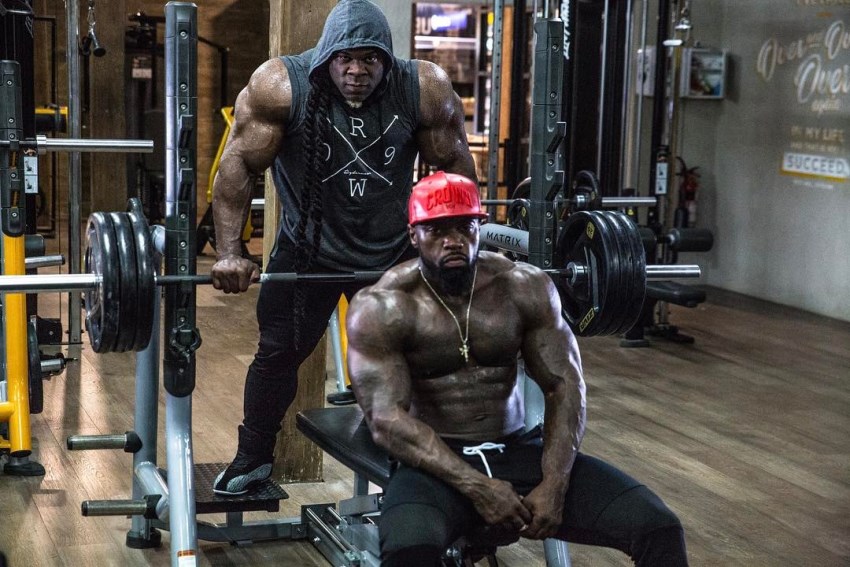 Mike Rashid sitting shirtless on a bench, preparing to do a heavy bench press, as Kai Greene spots him from behind
