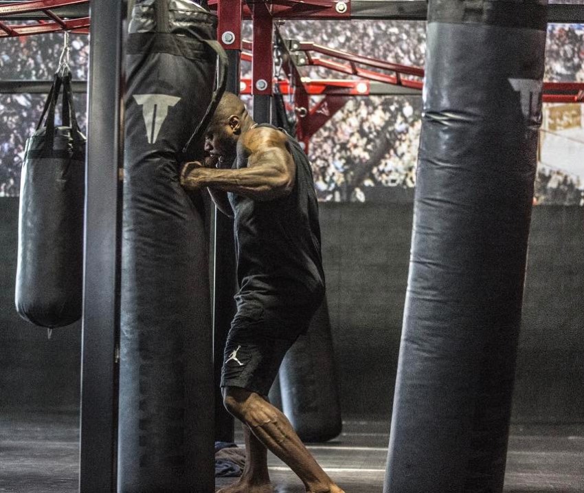 Mike Rashid wearing a black tank top and black shorts, punching a boxing back with his strong and muscular arms