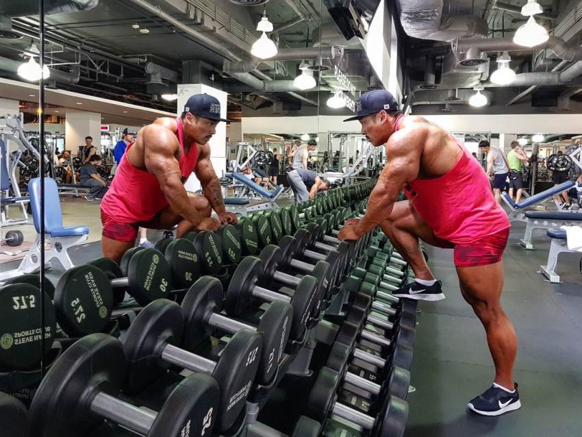 Nam Eun Cho looking at himself in the gym mirror, leaning against the dumbbell rack, looking massive and aesthetic