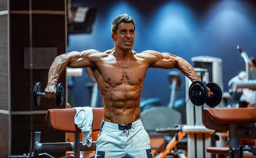 Nikolay Kuleshov training his side delts with dumbbells, being shirtless, looking ripped and muscular