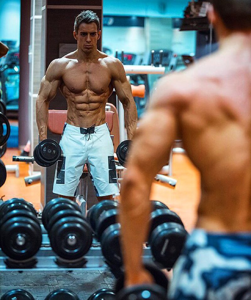 Nikolay Kuleshov standing by the gym mirror, looking at his ripped body as he performs an exercise with dumbbells