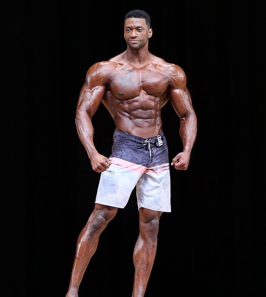 Raymont Edmonds posing on a Men's Physique stage, looking tall, muscular, and conditioned