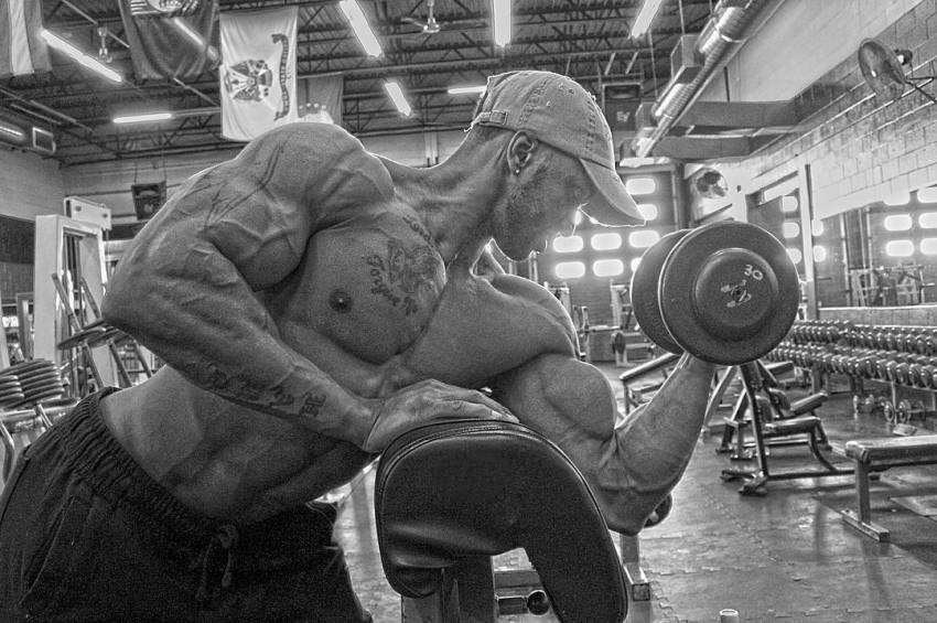 Raymont Edmonds being shirtless in a gym, doing dumbbell concentration curls for his biceps, looking riped and muscular