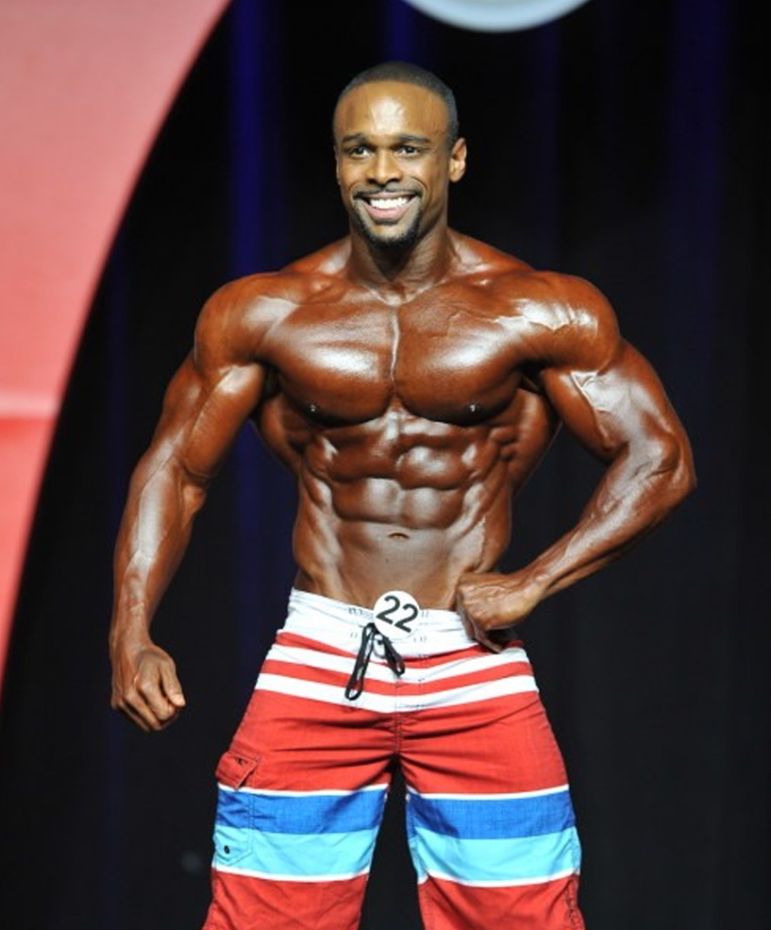 Ryan Hinton posing on the Mr. Olympia stage in red and blue shorts, displaying his ripped and muscular physique from the front